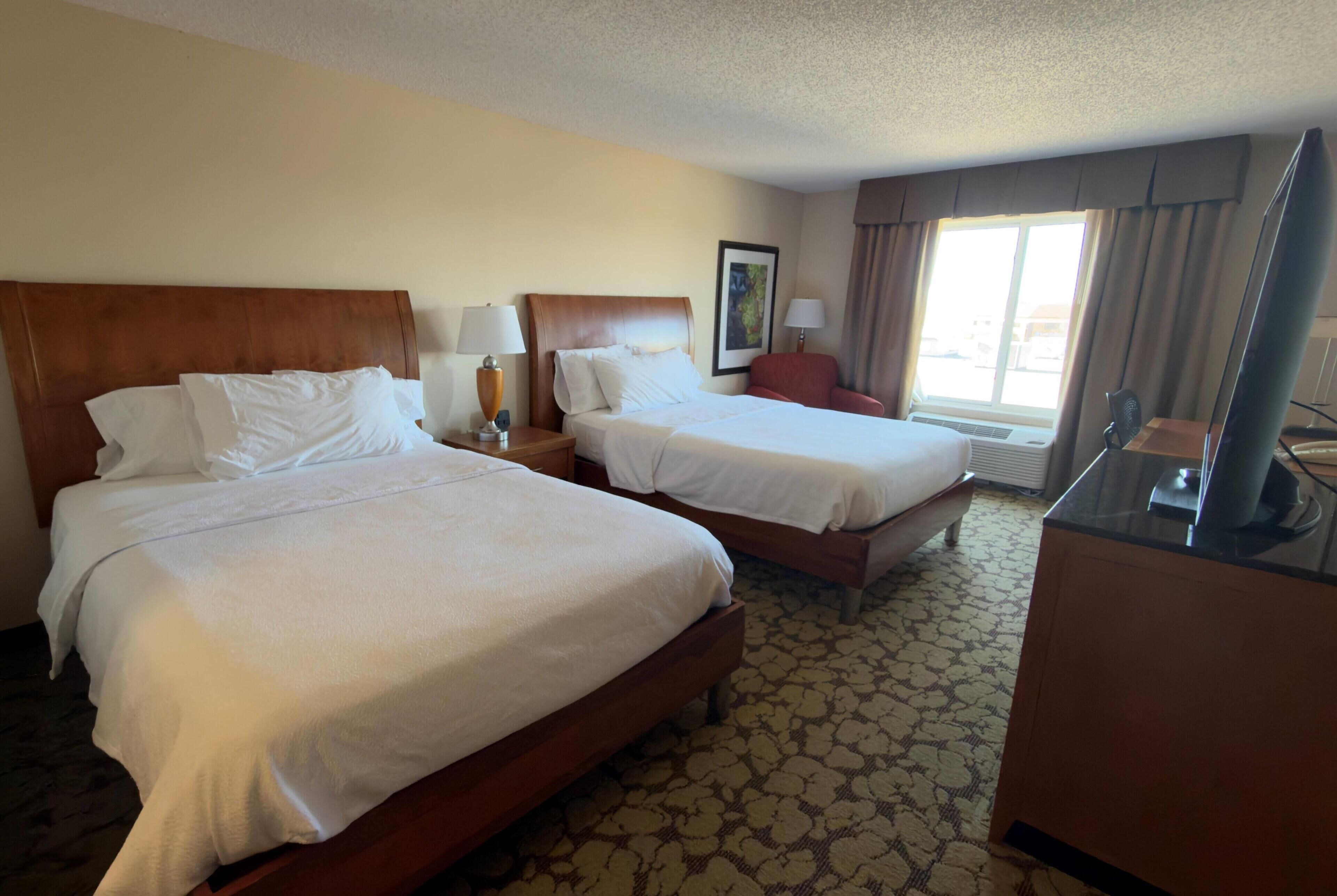 A hotel room with two beds, a desk, a chair, a TV, and a window with curtains.