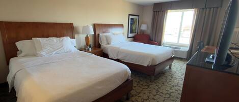 A hotel room with two beds, a desk, a chair, a TV, and a window with curtains.