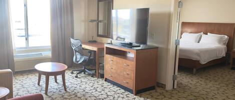 A hotel room with a bed, a desk with a TV, a chair, a small table, and a window with curtains.