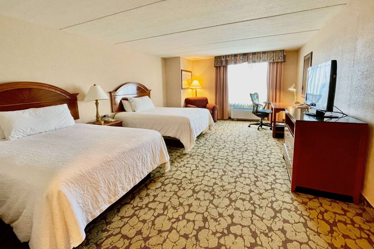 Quarto de hotel com duas camas, uma escrivaninha com cadeira, televisão e janela com cortinas.
