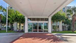 L’entrée d’un immeuble est agrémentée d’un chemin pavé de briques menant à des portes en verre, le tout étant entouré de verdure et de palmiers.