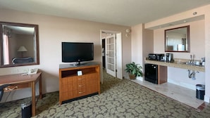 Une chambre d’hôtel avec un téléviseur posé sur un meuble en bois, un bureau avec un téléphone, une petite poubelle, une moquette à motifs, une kitchenette équipée d’un four à micro-ondes, d’une machine à café et d’un évier, ainsi qu’un miroir au-dessus de l’évier.