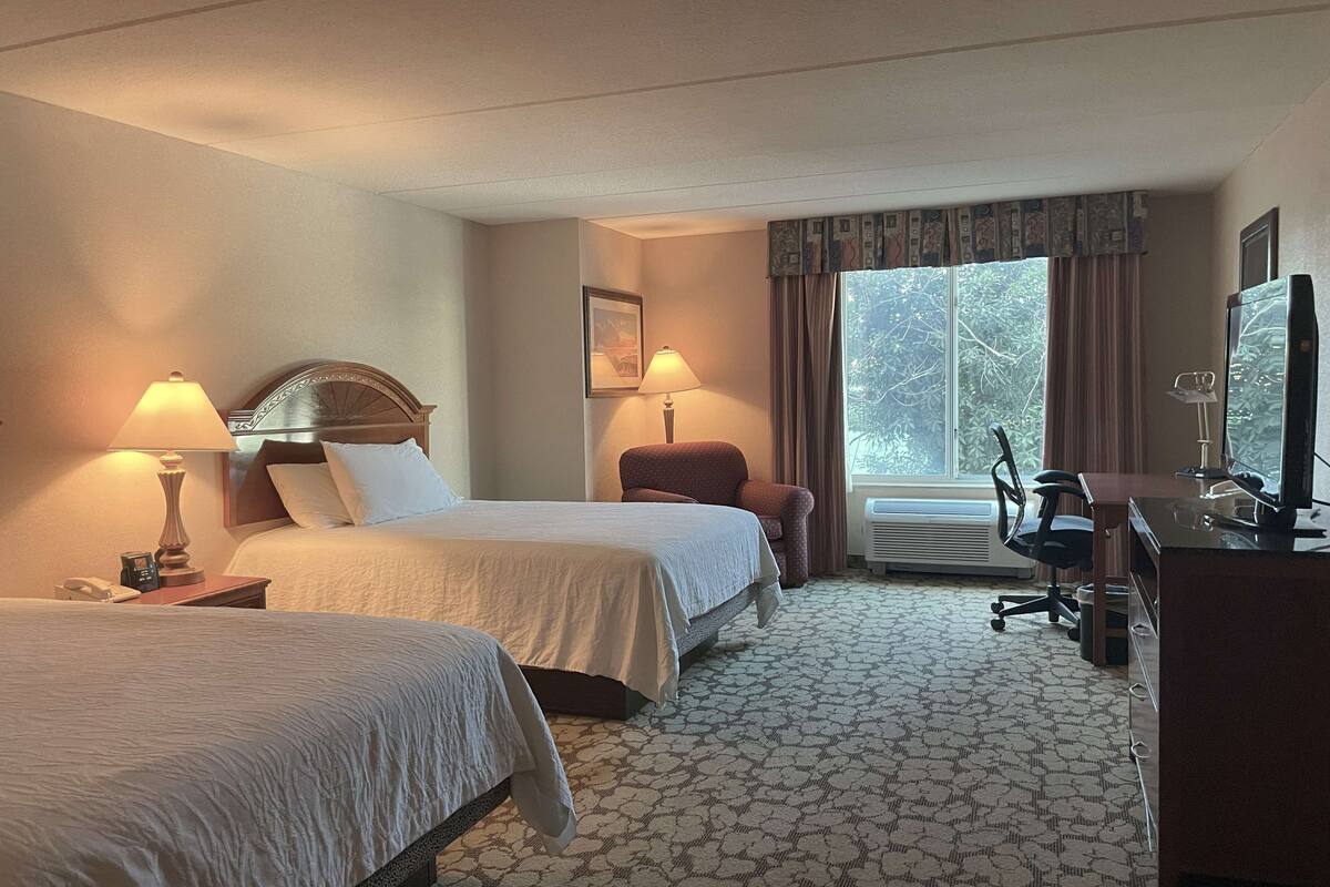 Quarto de hotel com duas camas, uma escrivaninha com computador, uma cadeira e uma janela com cortinas.