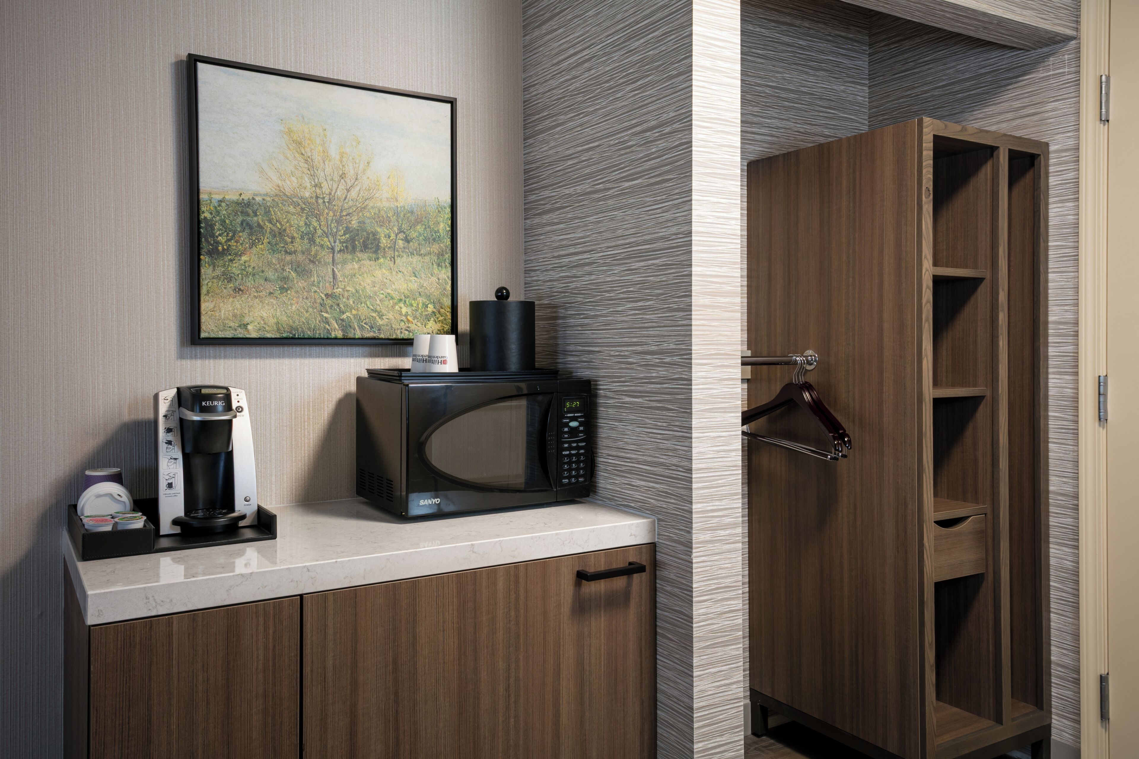 A kitchenette with a coffee maker, microwave, and a framed landscape painting on the wall.