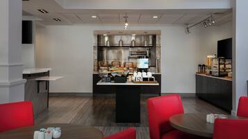 A modern hotel kitchen with a central island, stainless steel appliances, and a seating area with red chairs.