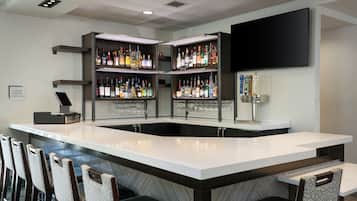 A bar area with a white countertop, multiple shelves of liquor bottles, a mounted TV, and bar stools.