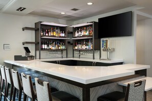 A bar area with a white countertop, multiple shelves of liquor bottles, a mounted TV, and bar stools. - Hilton Garden Inn Tri-Cities/Kennewick (Kennewick)
