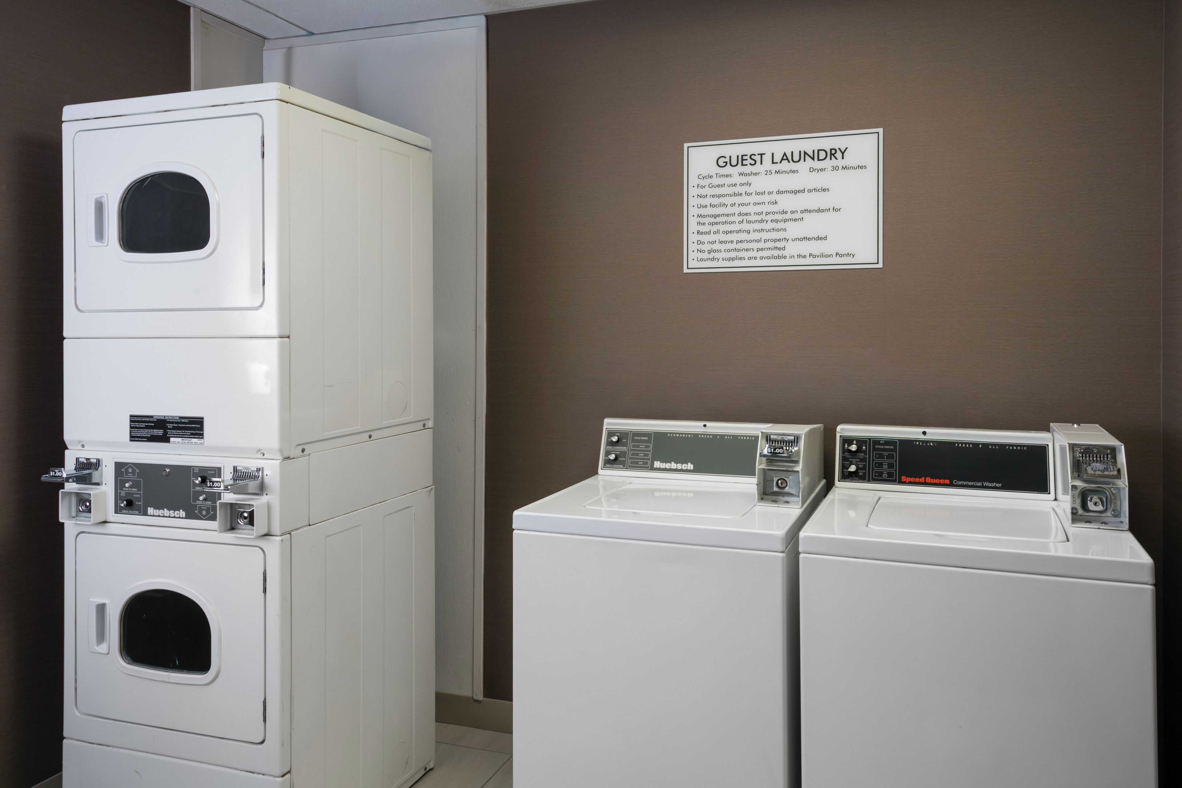 Een stapel van drie witte Huebsch wasmachines en drogers, met een bord erboven waarop staat 'GANGBARE WASSERIJ' en wascyclusduur.