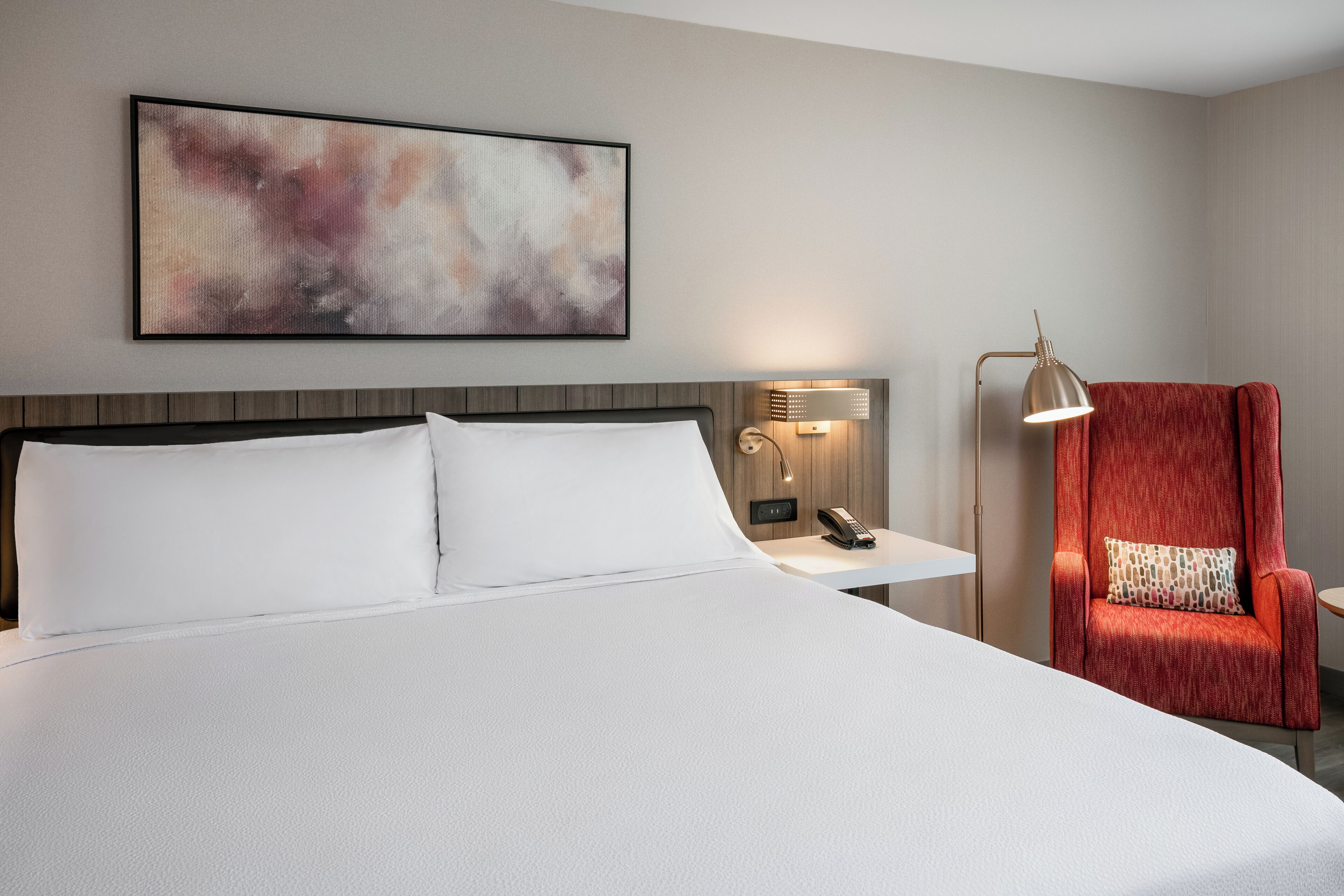 Hotellihuone, jossa on suuri sänky, punainen nojatuoli, yöpöytä lampun kanssa ja taulu seinällä.