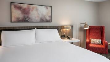 Hotellihuone, jossa on suuri sänky, punainen nojatuoli, yöpöytä lampun kanssa ja taulu seinällä.