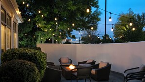 An outdoor seating area with wicker furniture and a fire feature, surrounded by a white wall and string lights.