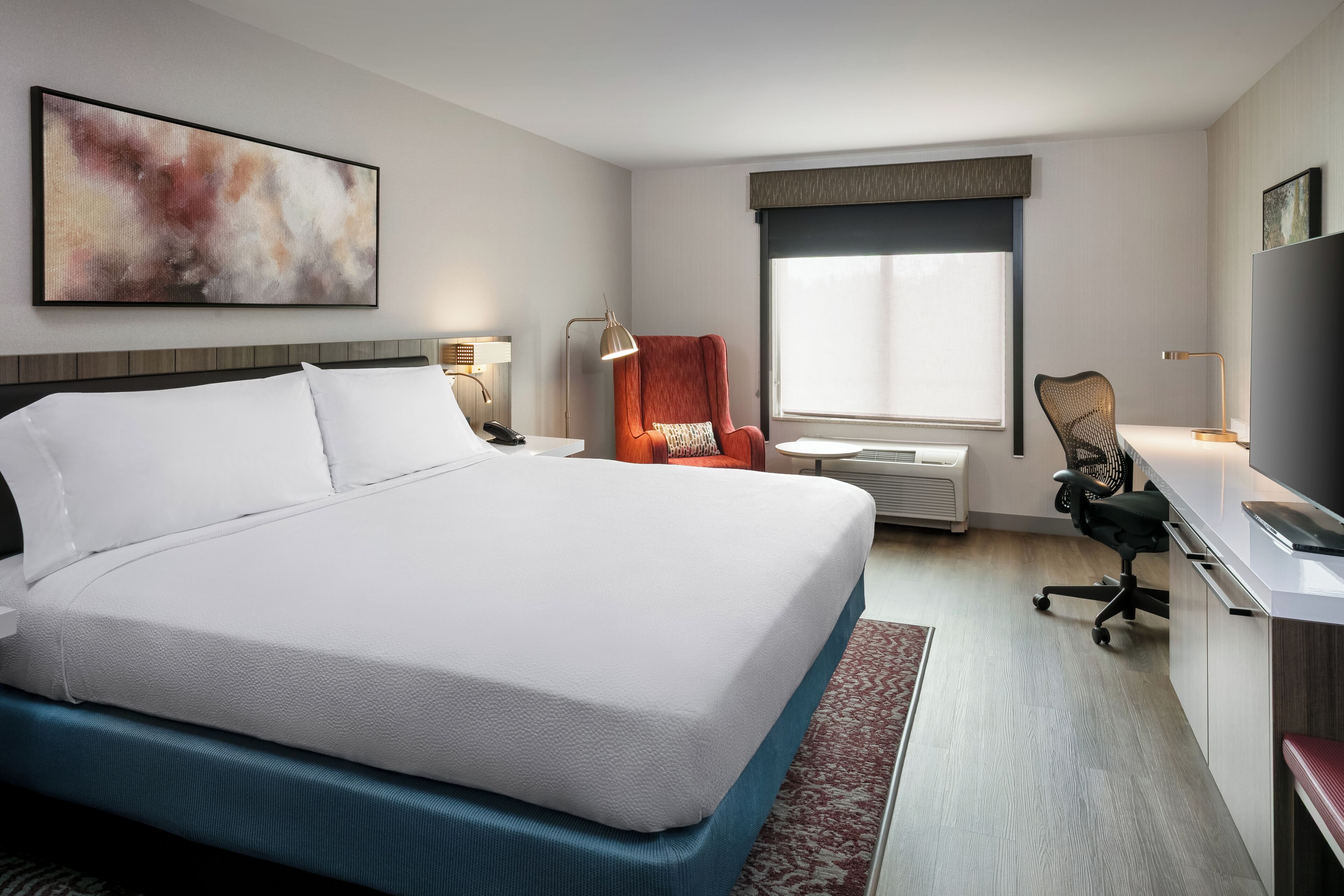 A hotel room with a large bed, a desk with a chair, a TV, and a window with blinds.