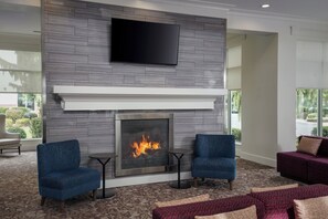 A modern living room with a fireplace, two blue armchairs, a red sofa, and a flat-screen TV mounted on the wall. - Hilton Garden Inn Tri-Cities/Kennewick (Kennewick)