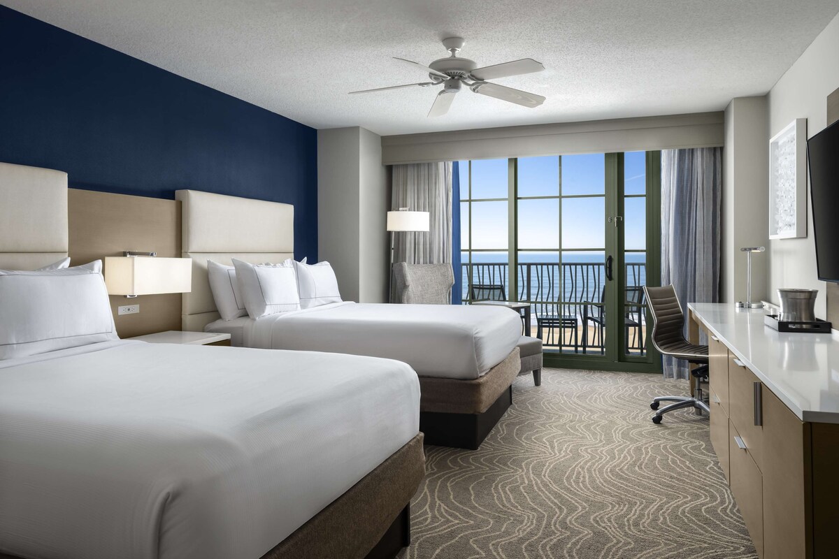 A hotel room with two beds, a desk, a chair, a ceiling fan, and a view of the ocean.