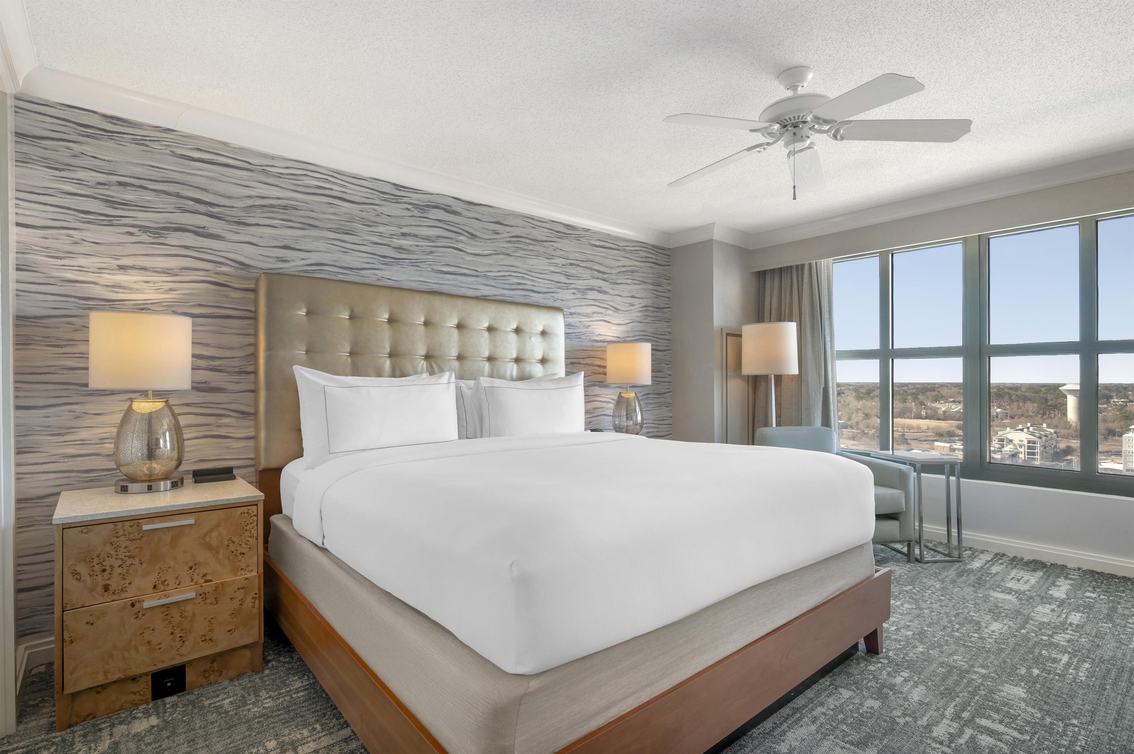 A hotel room with a large bed, two bedside lamps, a ceiling fan, and a view of the city through large windows.