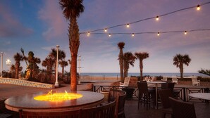 An outdoor seating area with tables and chairs, a fire pit, and string lights against a backdrop of the ocean and palm trees. - Hilton Virginia Beach Oceanfront (Virginia Beach)