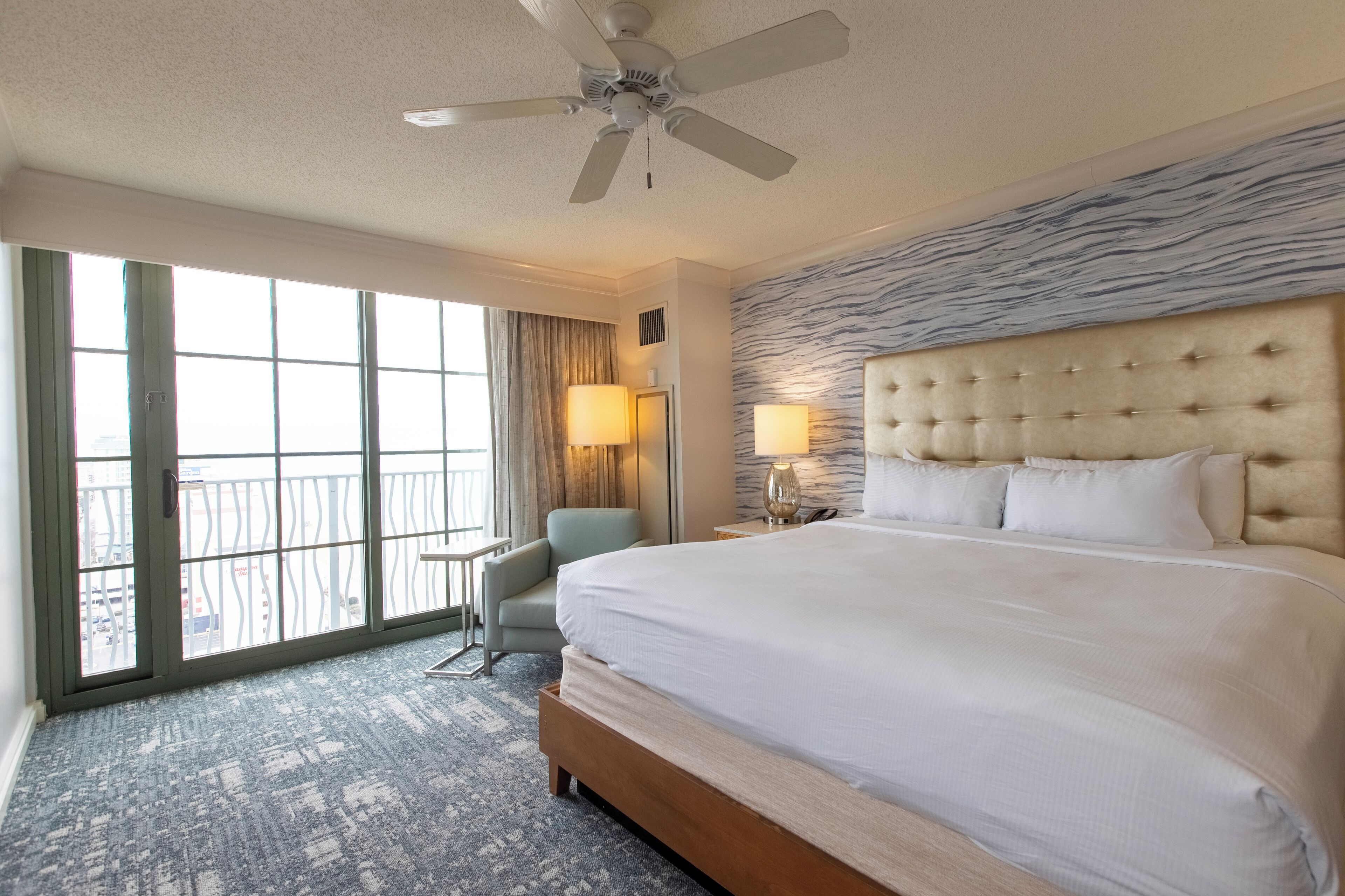 A hotel room with a large bed, a ceiling fan, a view through the window, and a chair.