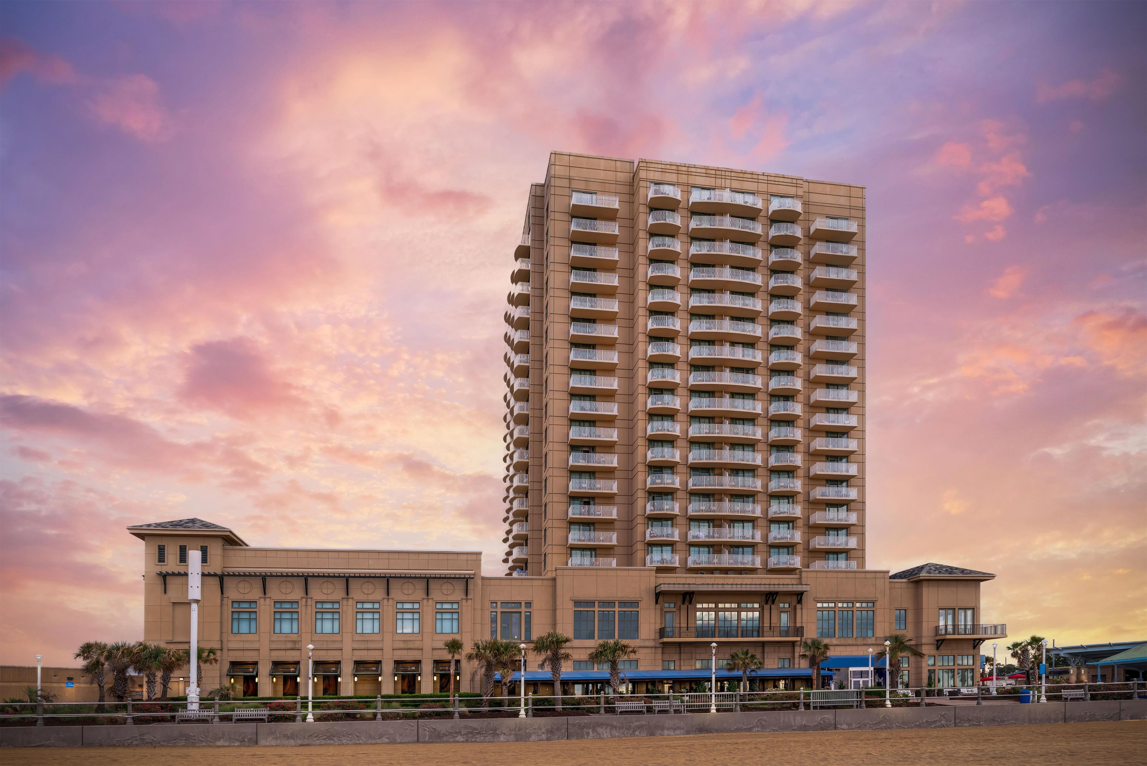 A high-rise hotel with multiple balconies, a central entrance, and a landscaped area with palm trees.