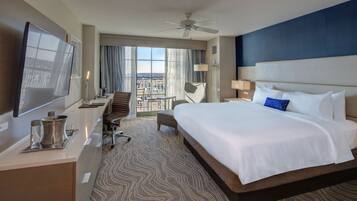 A hotel room with a large bed, a desk with a flat-screen TV, a chair, a balcony with a view, and a ceiling fan.