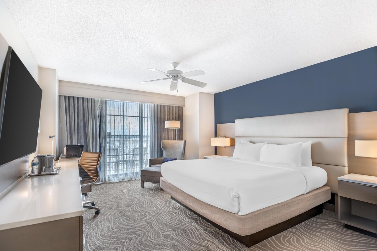 A hotel room with a large bed, a desk with a chair, a television, and a ceiling fan.