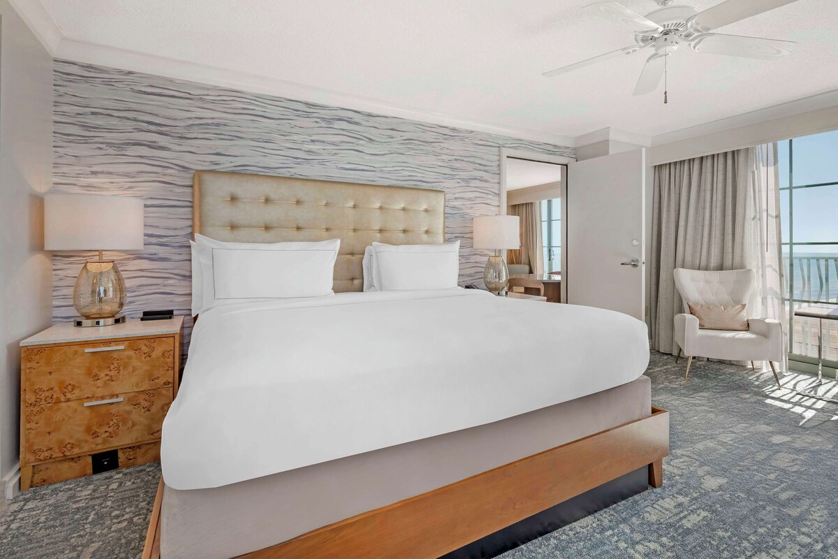 A large bed with a tufted headboard, two bedside tables with lamps, a chair, and a view of the outdoors.