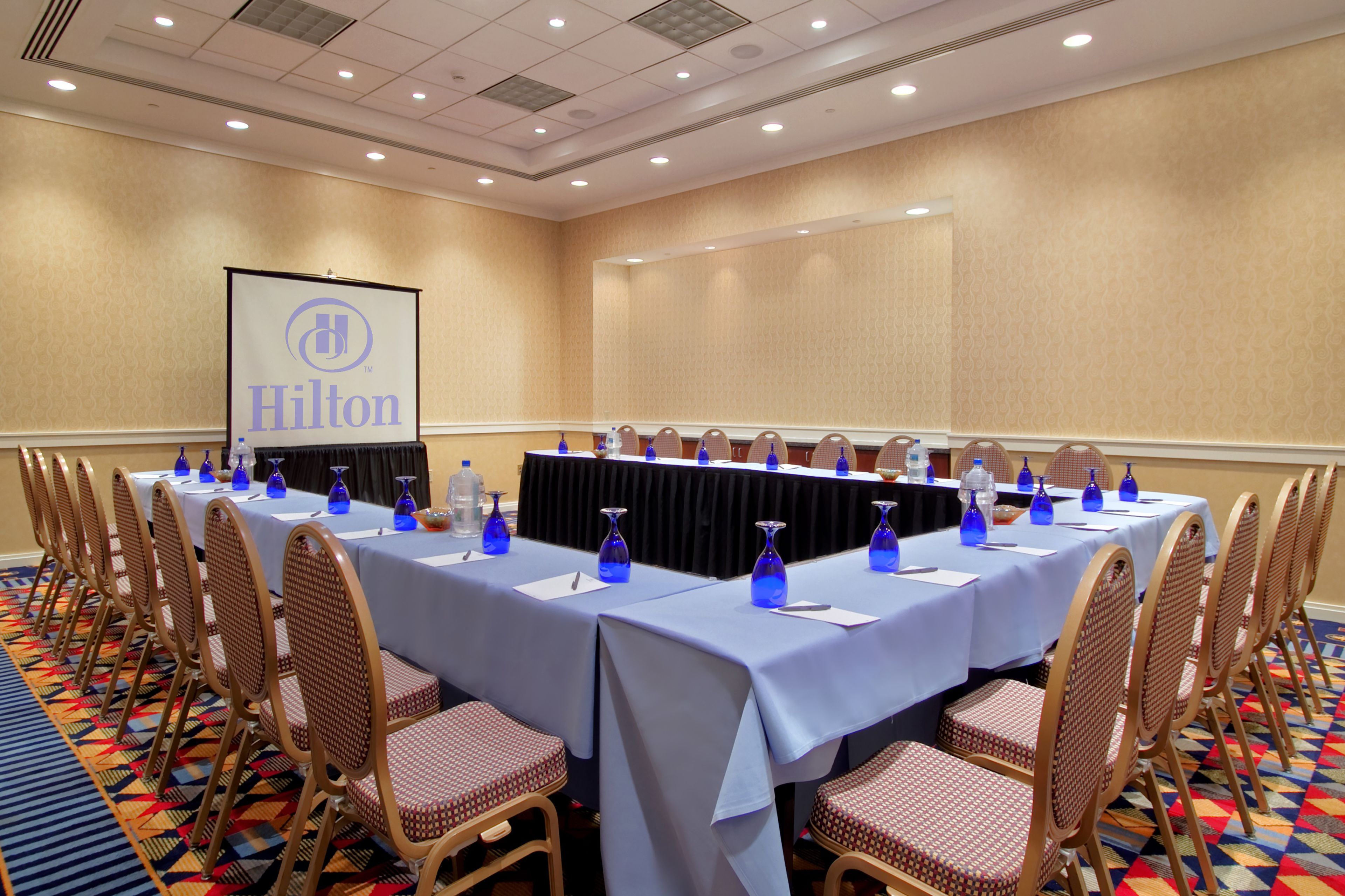 A conference room with a long table, chairs, and a projector screen displaying 'Hilton'.