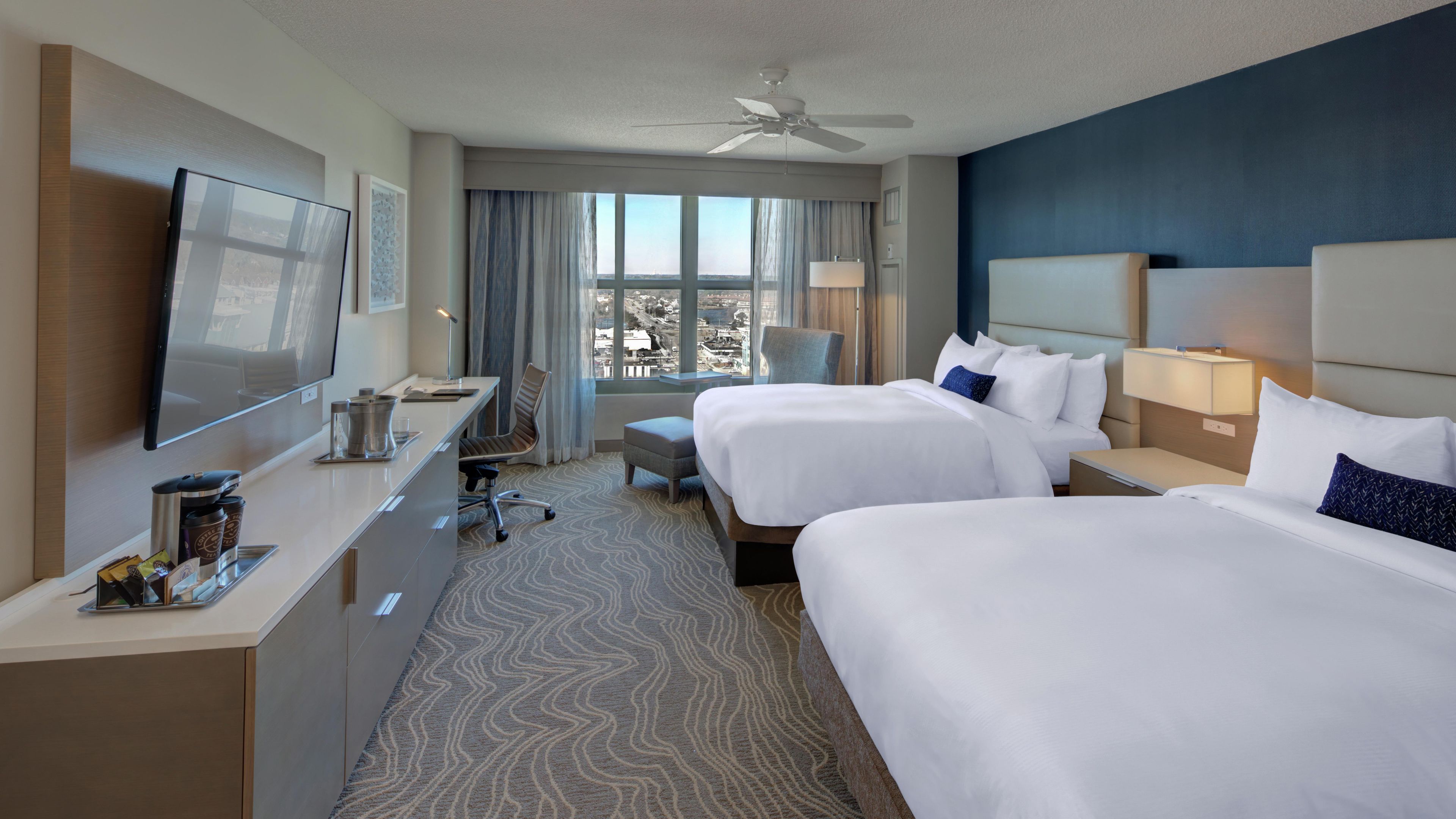 A hotel room with two beds, a desk, a TV, and a view of the city.