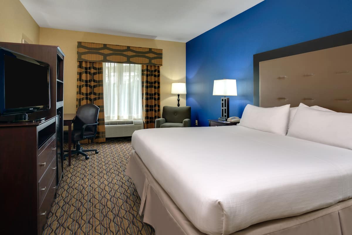 A hotel room with a large bed, a desk with a computer, a chair, a TV, and a window with curtains.