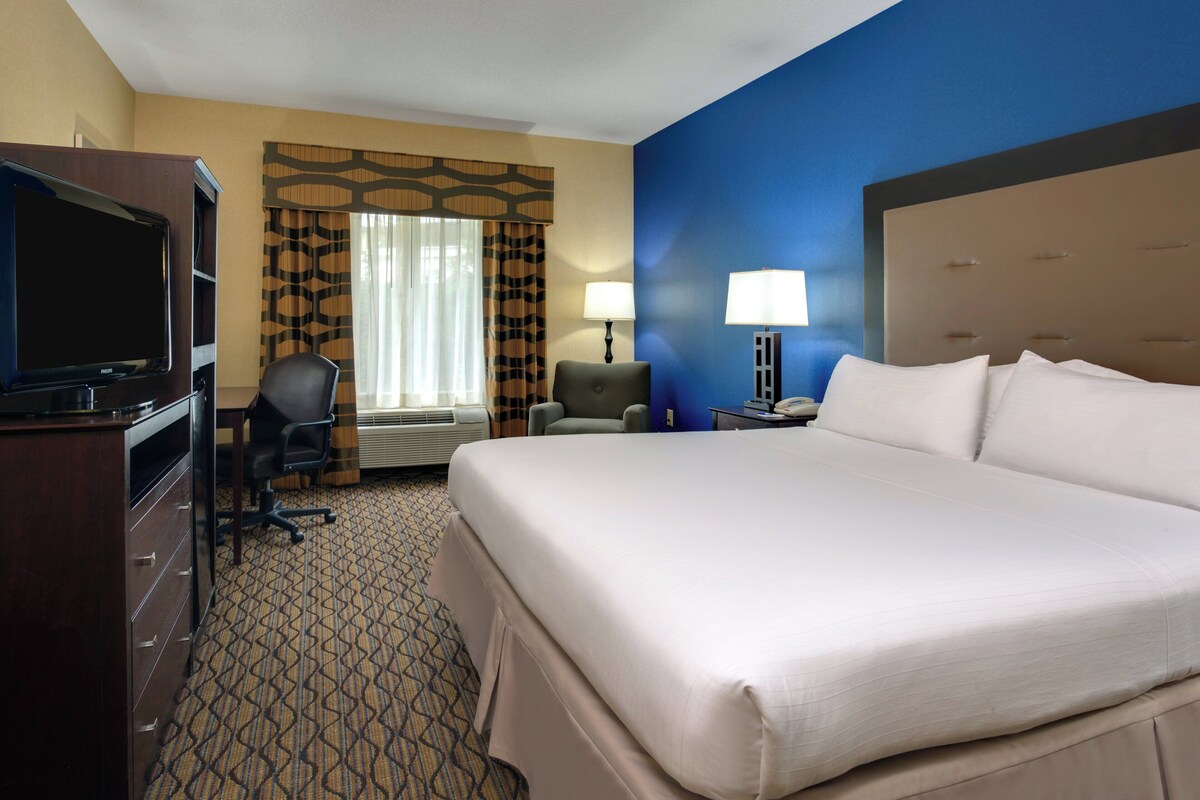 A hotel room with a large bed, a desk with a computer, a television, and a window with curtains.