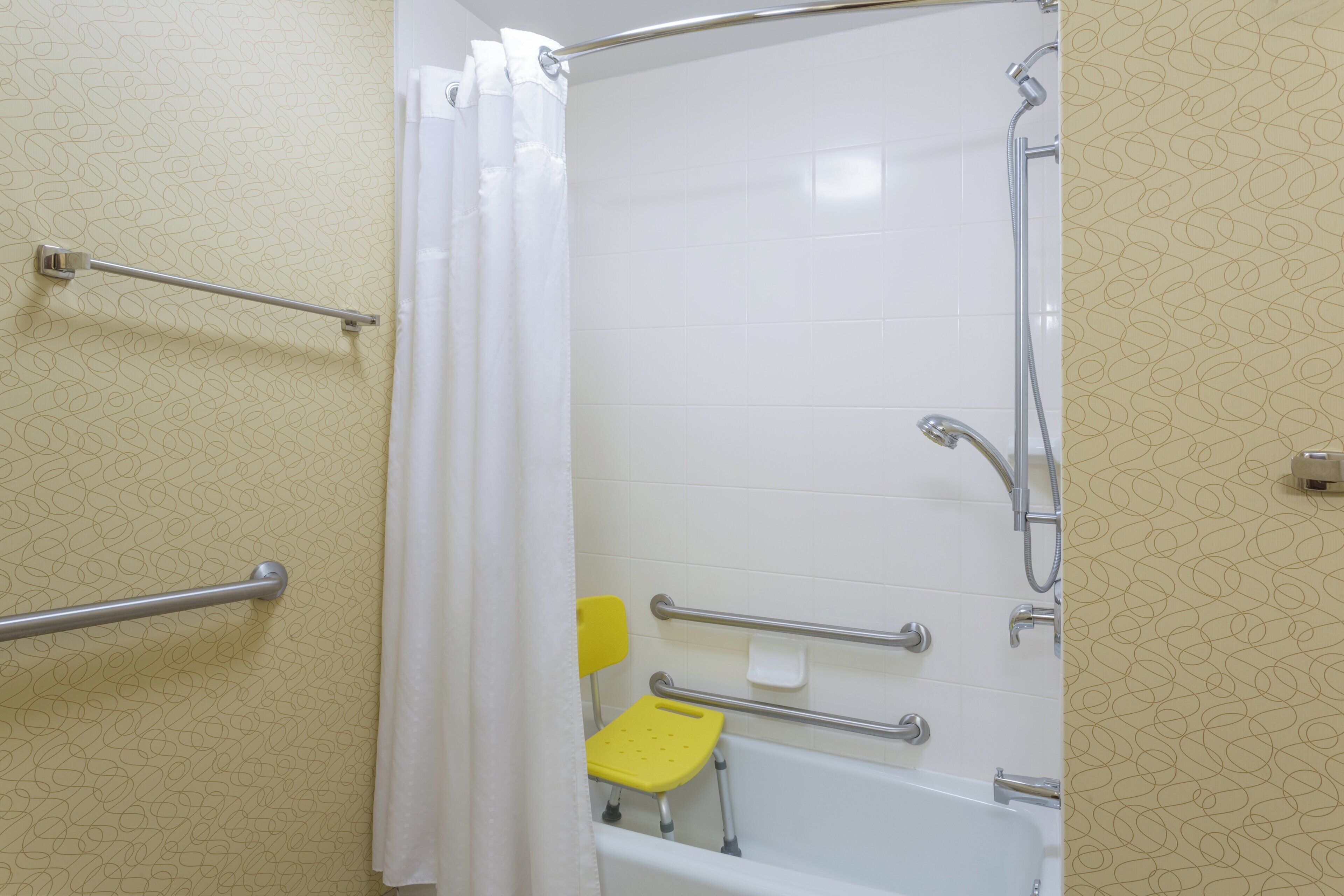 A bathroom with a shower, grab bars, a yellow seat, and a white bathtub.
