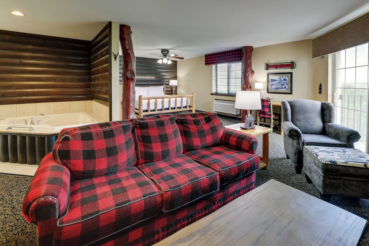 A hotel room with a checkered sofa, a gray armchair, a wooden coffee table, a large window with curtains, a ceiling fan, a bed with a wooden headboard, and a radiator.