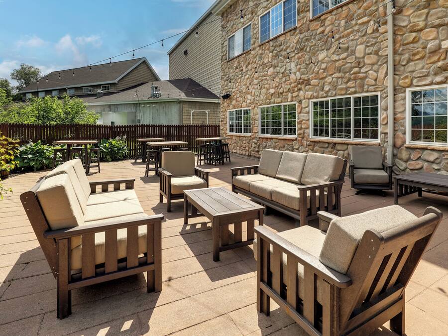 An outdoor patio area with wooden furniture, including sofas, chairs, and tables, set against a stone building with large windows.