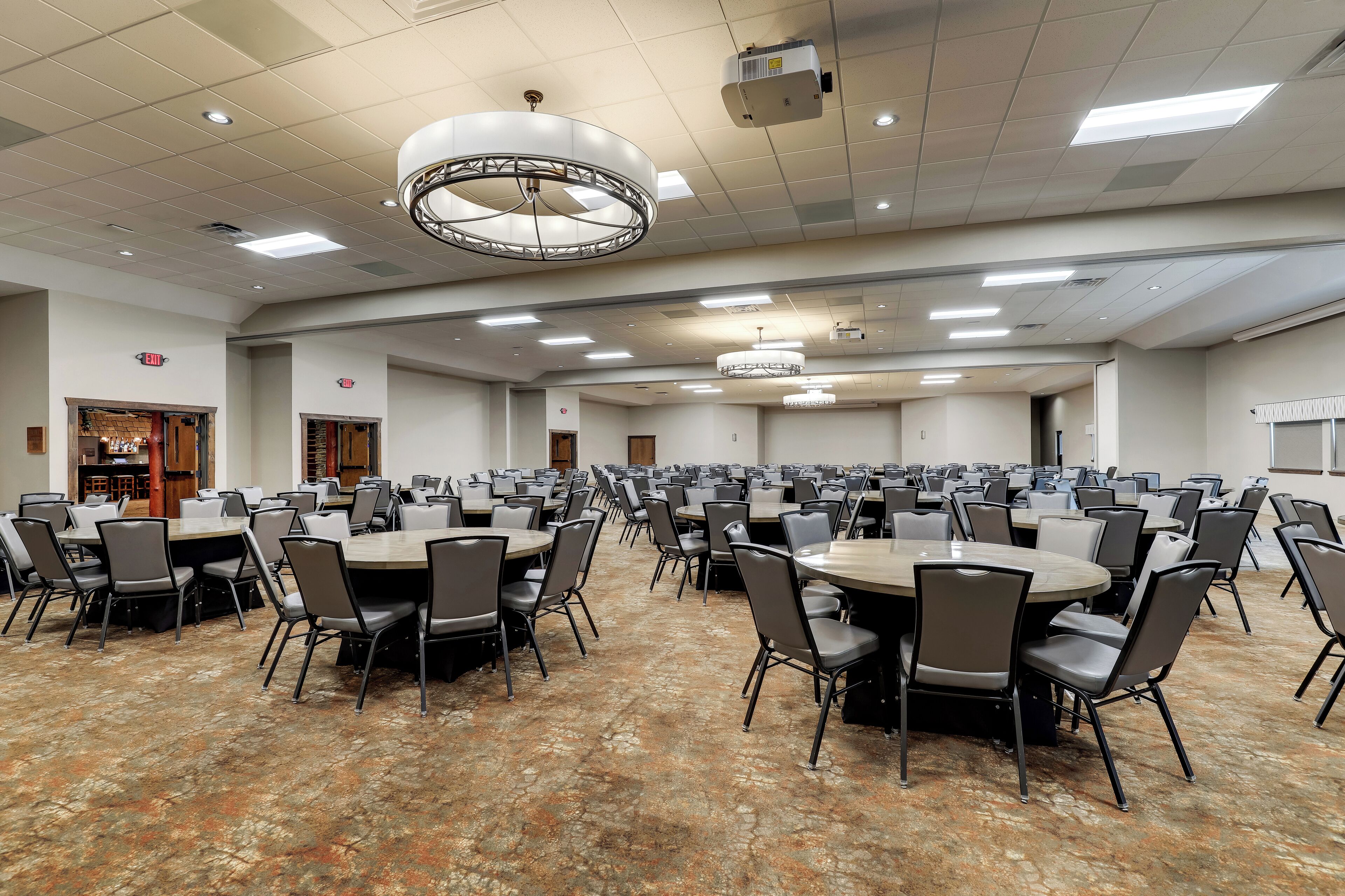 A large conference room with multiple round tables and chairs, ceiling lights, and an exit sign.