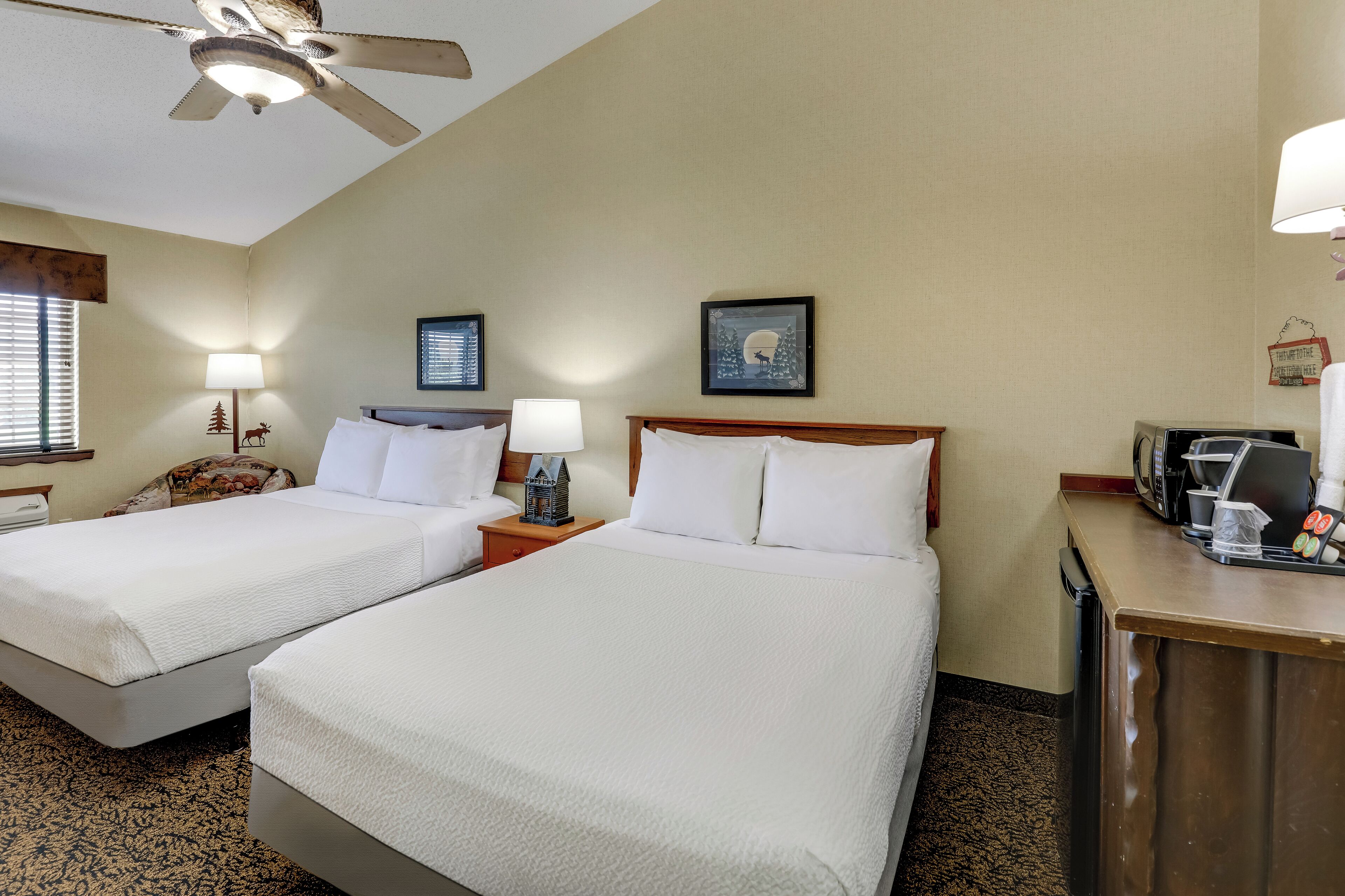 A hotel room with two beds, a ceiling fan, a desk with a coffee maker, and framed pictures on the walls.