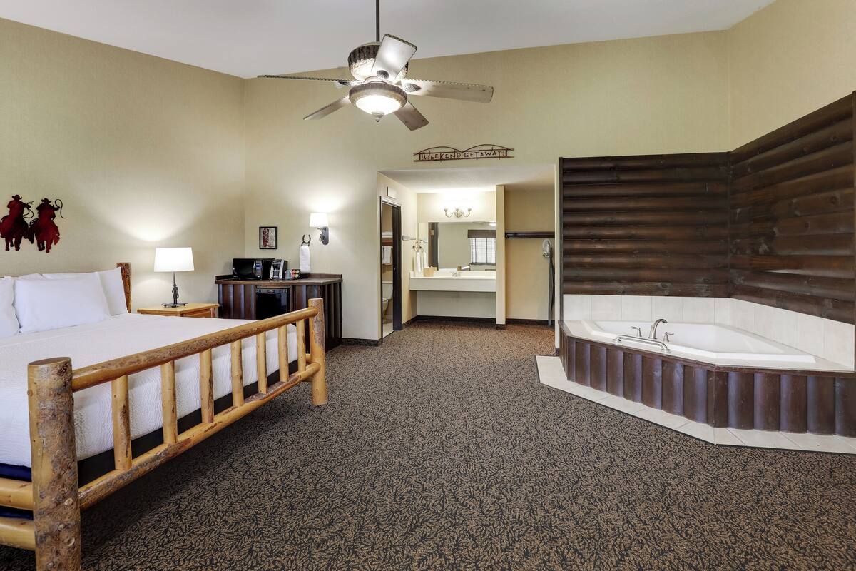 A hotel room with a bed, a wooden headboard, a large bathtub, a ceiling fan, a desk with a lamp, and a mirror.
