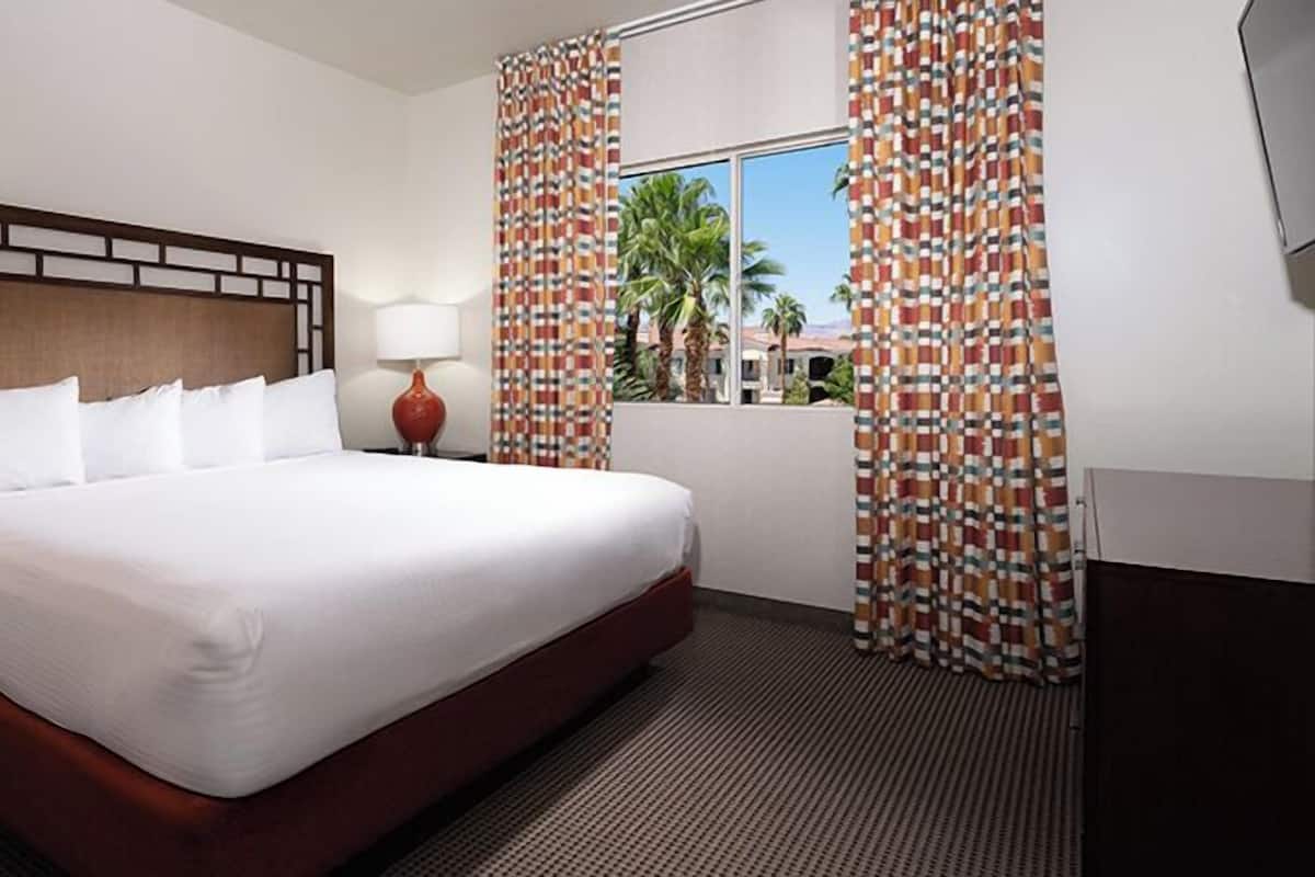 A hotel room with a large bed, two bedside lamps, a window with curtains, and a view of palm trees.
