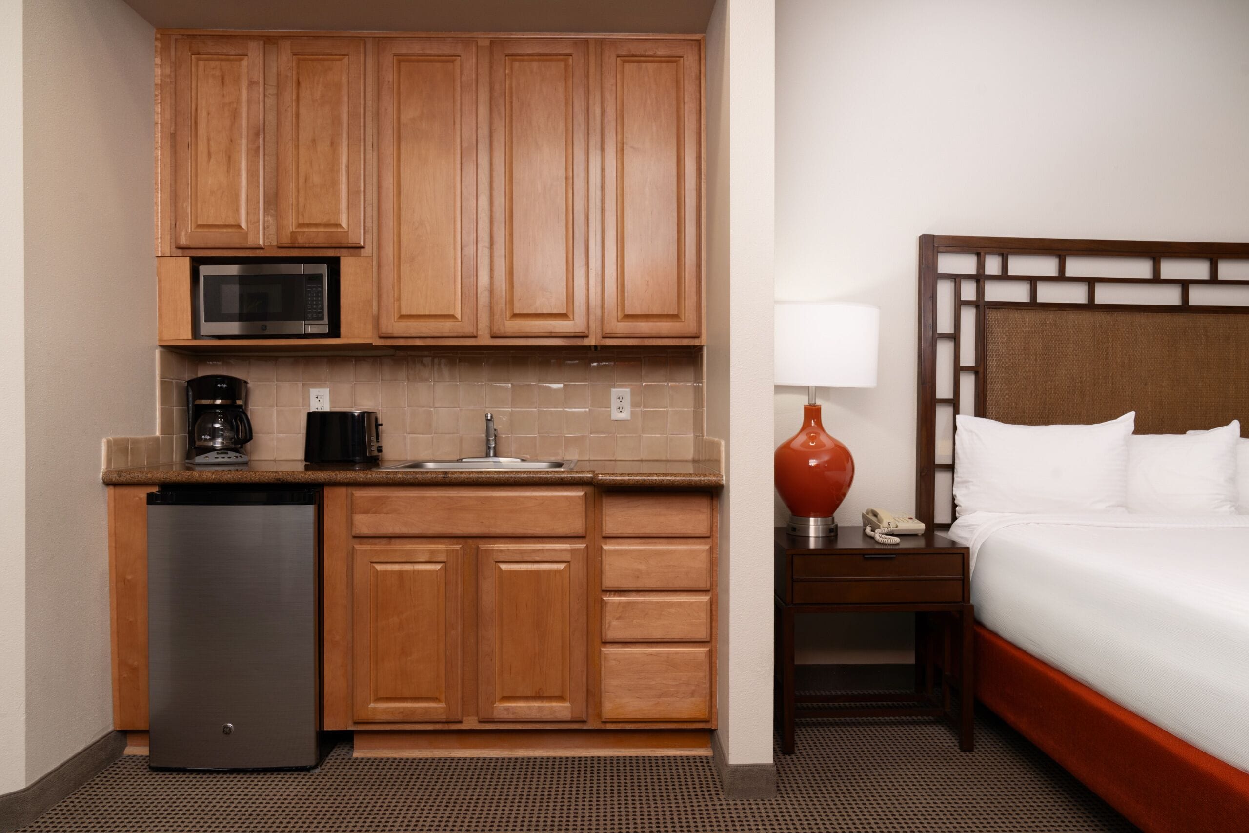 Habitación de hotel compacta con kitchenette, que incluye armarios de madera, microondas, cafetera y un refrigerador pequeño. Cuenta con una cama con ropa blanca y una mesita de noche con lámpara y teléfono.