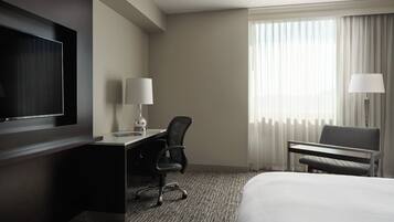 Habitación de hotel con cama, escritorio, silla, televisor y ventana con cortinas.