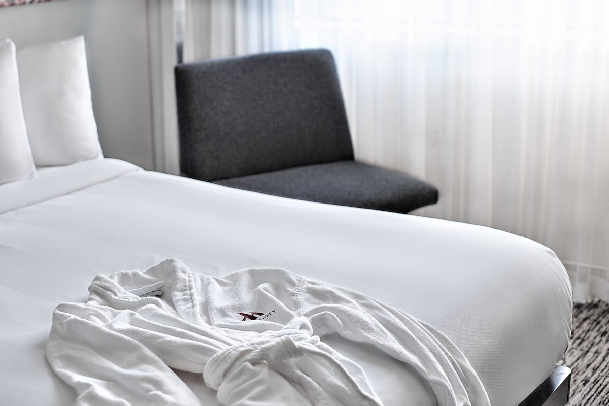 A hotel room with a neatly made bed, a grey armchair, and a window with sheer curtains.