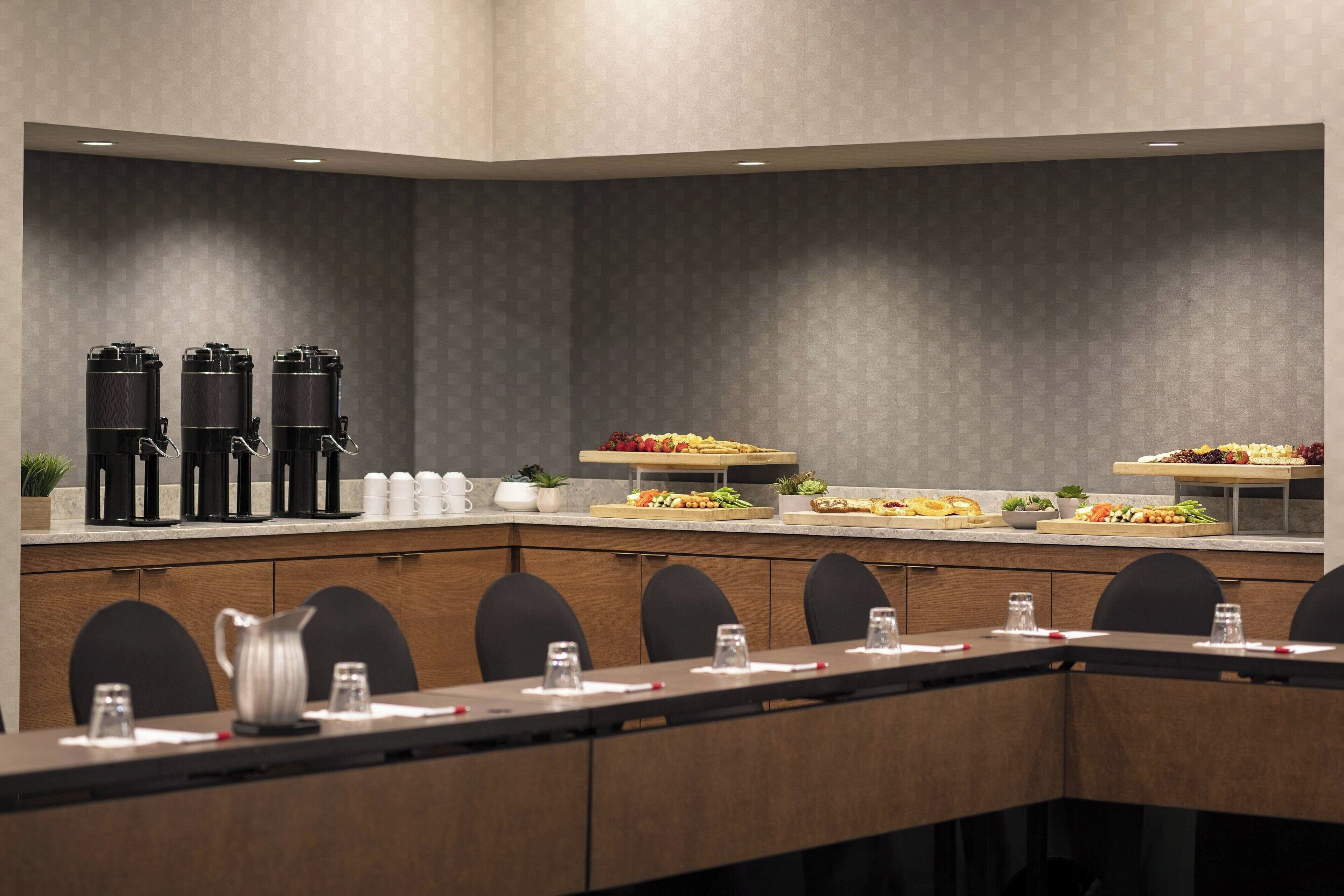 A modern conference room with a long table, black chairs, a coffee station, and a buffet with various food items.