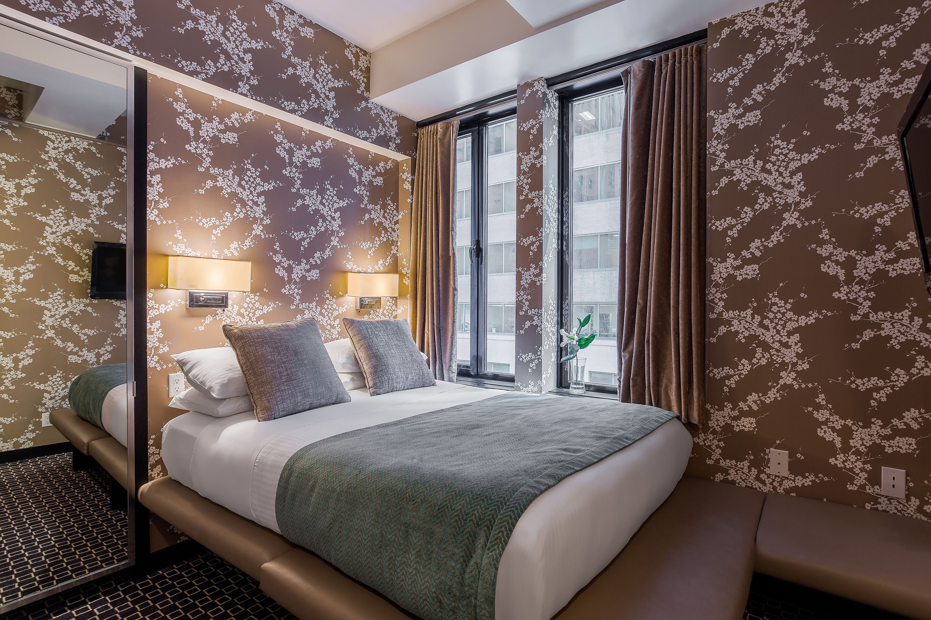 Une chambre d’hôtel avec un lit, un miroir, une fenêtre avec des rideaux et un mur recouvert d’un papier peint à motifs floraux.