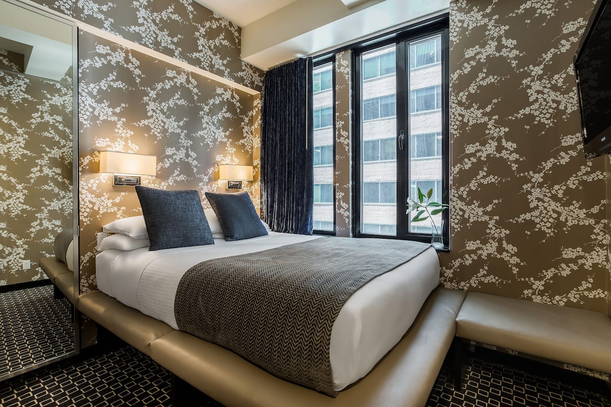 Camera d'albergo con un letto grande, due lampade ai lati del letto, una panca e una finestra con vista su un edificio.