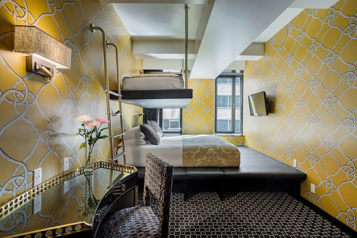Camera d'albergo compatta con un letto a castello, una scrivania con sedia, un tavolino e un vaso decorativo con fiori.