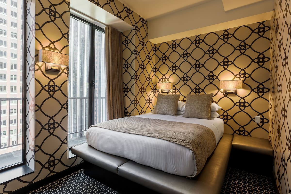 Quarto de hotel com cama, abajures, janela e papel de parede estampado.
