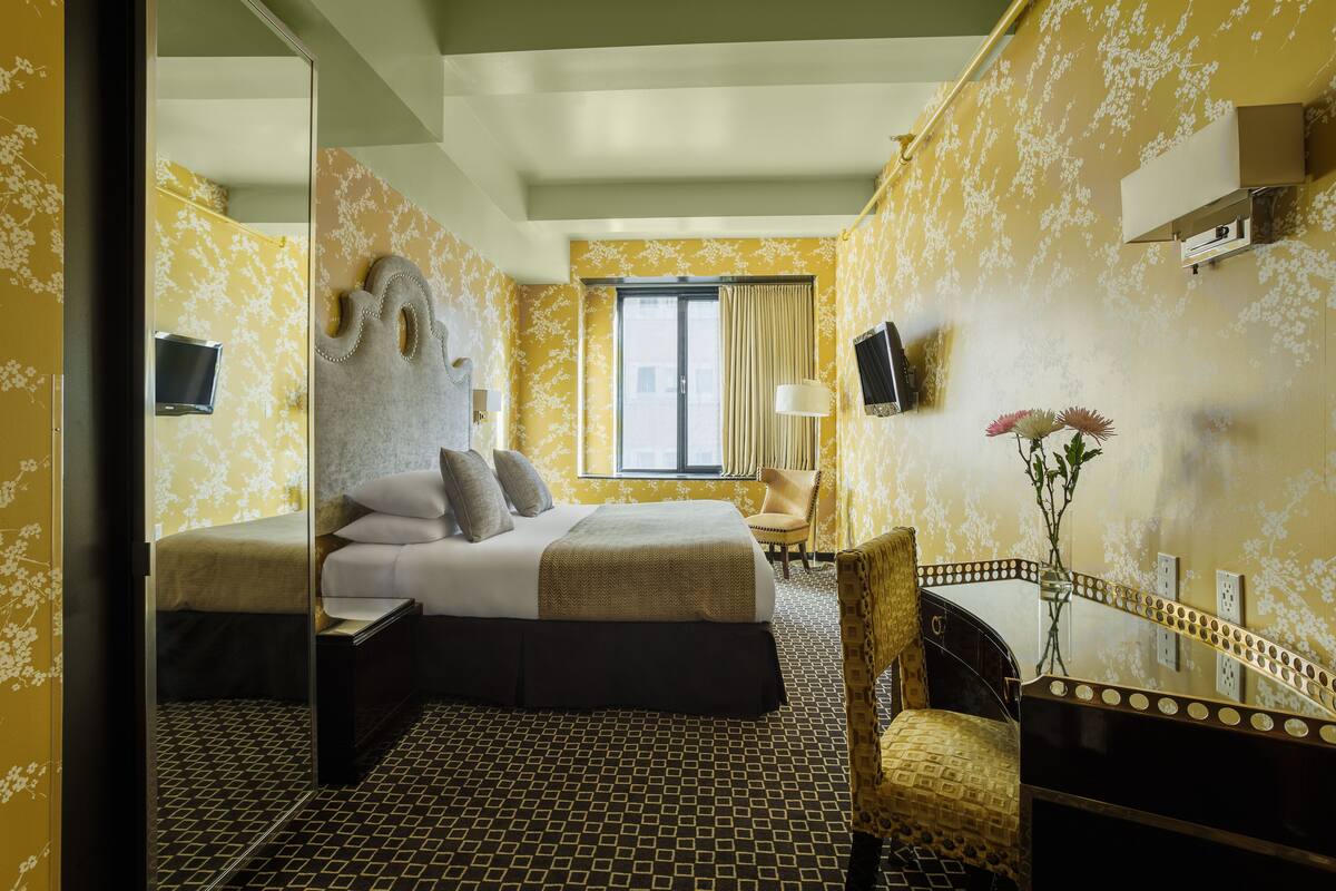 Una camera d'albergo con un letto, una scrivania, una sedia, uno specchio, una finestra e una TV a parete.