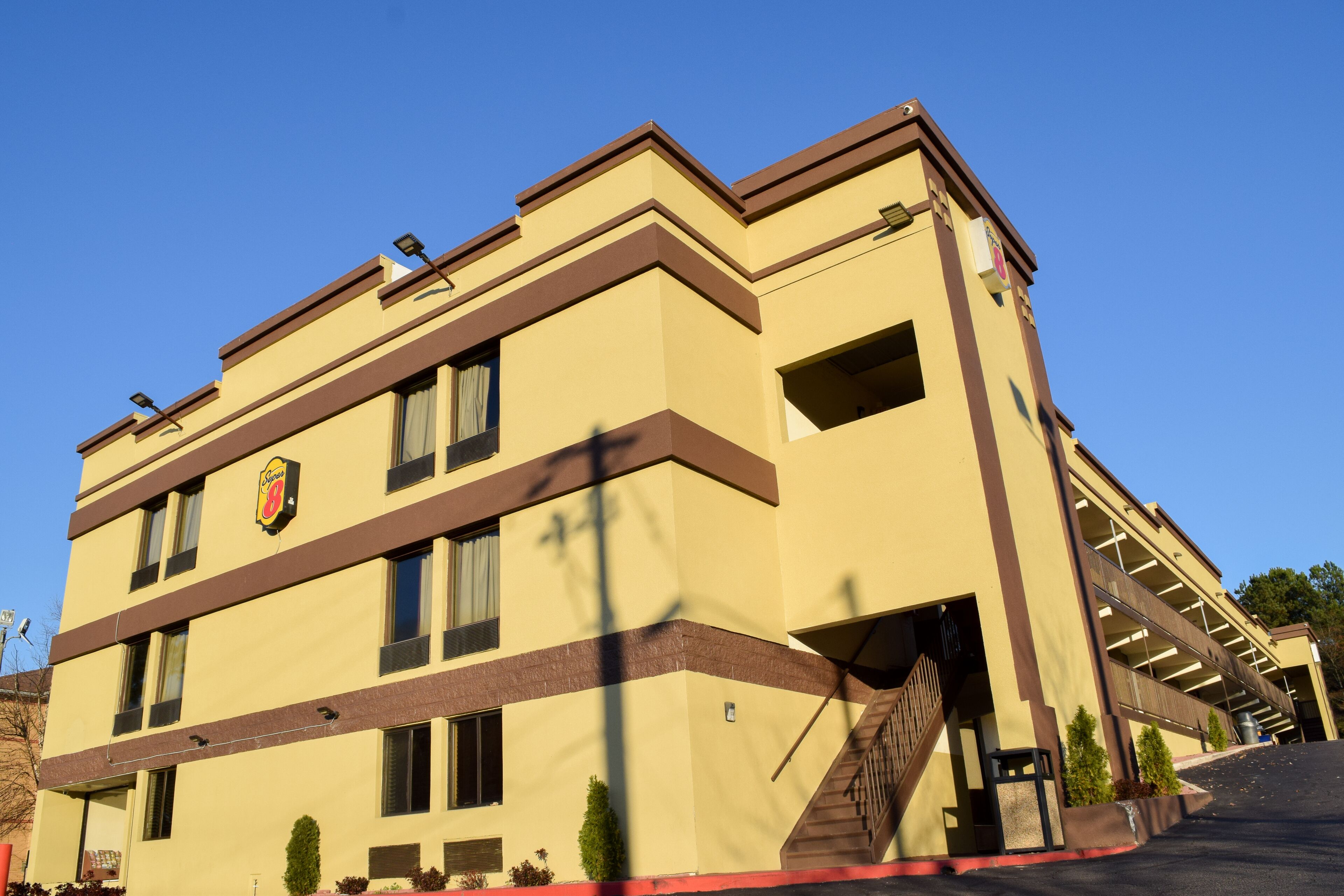 A multi-story hotel with a beige facade, brown trim, and a visible logo. It features a staircase leading to the entrance and a parking area in front.