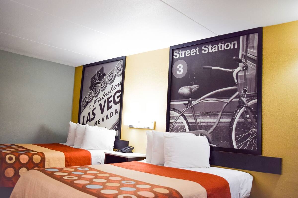 A hotel room with two beds, a nightstand, a lamp, and two posters on the wall.