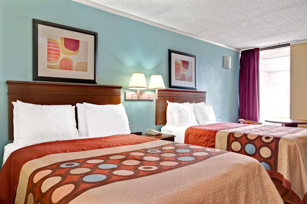 A hotel room with two beds, a nightstand with a lamp, and artwork on the walls.