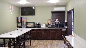 Een kleine, goed verlichte kamer met een witte tafel en stoelen, een aan de muur bevestigde televisie en een kitchenette met apparatuur en een koffieapparaat.