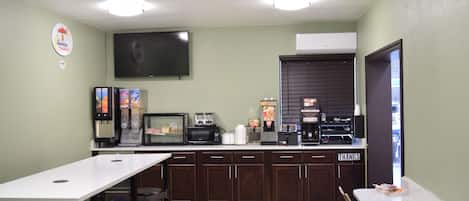 Una habitación pequeña y bien iluminada con una mesa y sillas blancas, un televisor montado en la pared y una zona de cocina con electrodomésticos y una cafetera.